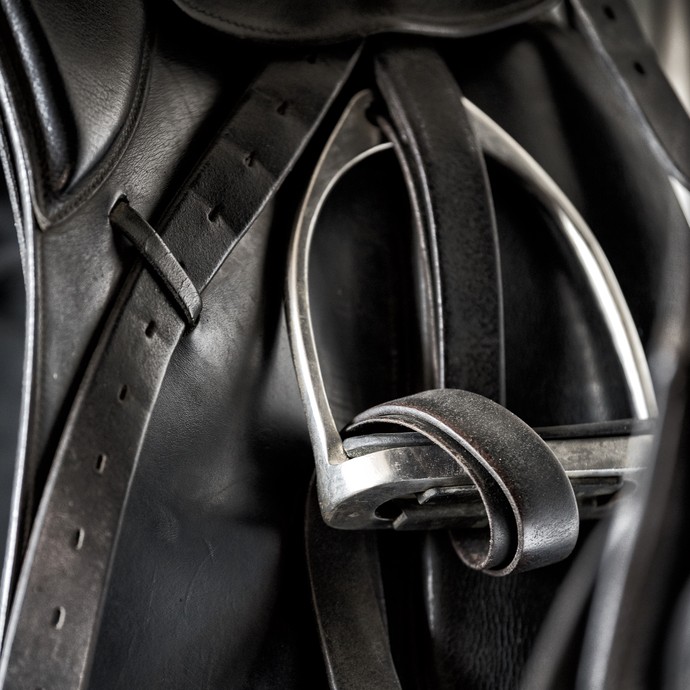 Detailfoto eines Steigbügels an einem schwarzen Dressursattel. Ein schwarzer Lederriemen ist durch den Steigbügel hindurch gezogen. (vergrößerte Bildansicht wird geöffnet)