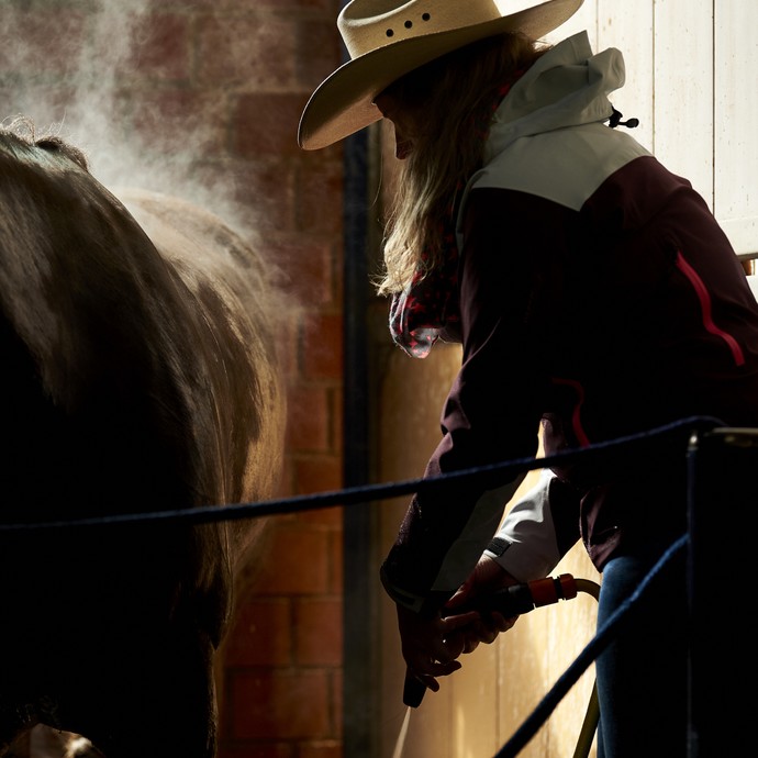 Eine Frau mit Cowboyhut betätigt einen Wasserschlauch. Neben ihr steht ein vom Schweiß dampfendes Pferd im Gegenlicht. (öffnet vergrößerte Bildansicht)