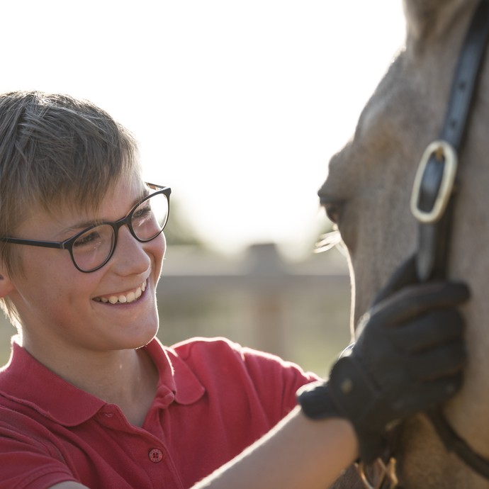 Ein blonder Junge mit rotem Poloshirt steht lachend einem hellen Pferd gegenüber. Er hält das Tier am Halfter und blickt ihm in die Augen. (vergrößerte Bildansicht wird geöffnet)