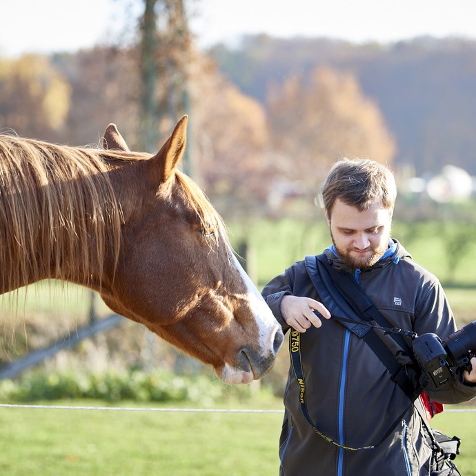 Ein braunes Pferd schnuppert an einem jungen Mann. Er hält eine Foto-Kamera in den Händen. Er ist ein Fotograf. (vergrößerte Bildansicht wird geöffnet)
