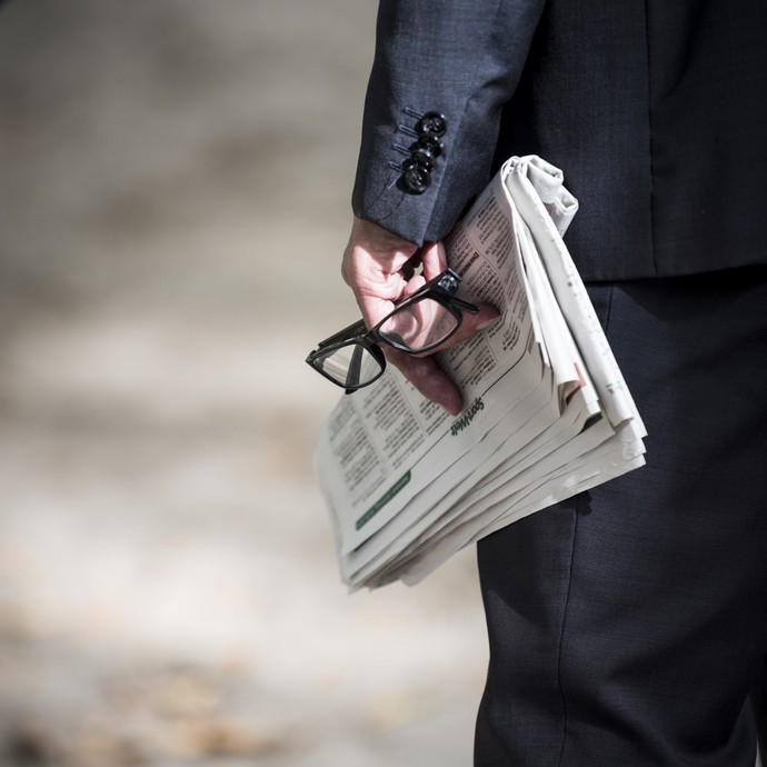 Ein Mann im Anzug hält eine Zeitung und eine Lesebrille in der Hand. In der Zeitung stehen die Startlisten eines Pferderennens. (vergrößerte Bildansicht wird geöffnet)