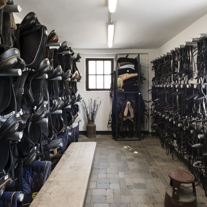 Innenraum einer Sattelkammer. An der linken Wand hängen schwarze Sättel in vier Reihen übereinander. rechts hängen Trensen und sonstifer Reitbedarf. (vergrößerte Bildansicht wird geöffnet)
