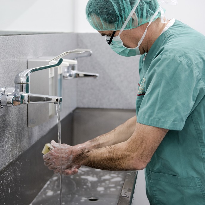 Ein Mann in OP-Kleidung wäscht seine Hände an einem großen Waschbecken aus Edelstahl. Er trägt einen grünen Kittel, Mundschutz und ein Haarnetz. (vergrößerte Bildansicht wird geöffnet)