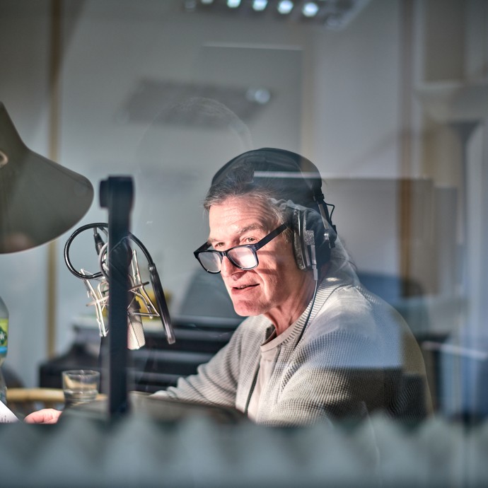 Der Sprecher Holger Löwenberg sitzt vor einem Mikrofon. Er trägt Kopfhörer und eine Brille. 
Er liest einen Text vor. (vergrößerte Bildansicht wird geöffnet)