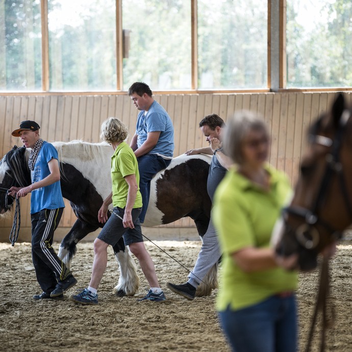 Teilnehmer einer Reittherapiestunde und zwei Frauen bewegen sich mit ihren Pferden durch eine Reithalle. (vergrößerte Bildansicht wird geöffnet)