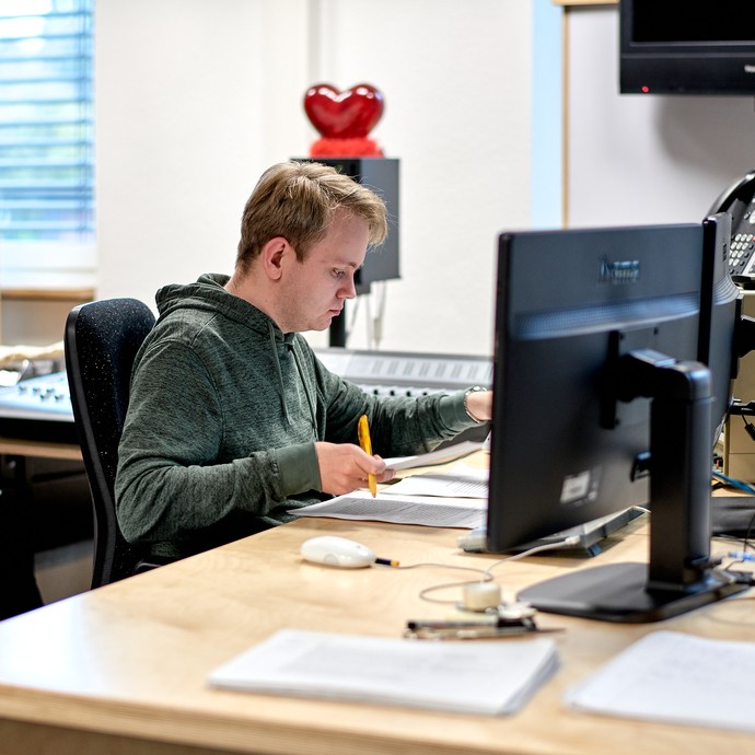 Ein junger Mann sitzt an einem Tisch in einem Ton-Studio.
Er arbeitet mit Papieren. Vor ihm stehen Bildschirme und ein Telefon. (vergrößerte Bildansicht wird geöffnet)