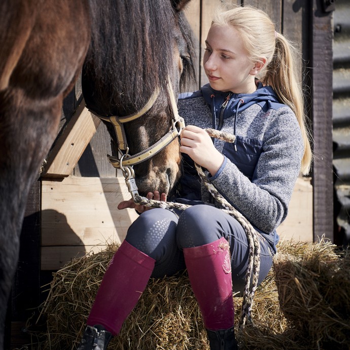 Ein blondes Mädchen sitzt auf einem Strohballen. Ein braunes Pferd neigt seinen Kopf zu ihr herunter, sie streichelt das Tier und lächelt. (vergrößerte Bildansicht wird geöffnet)