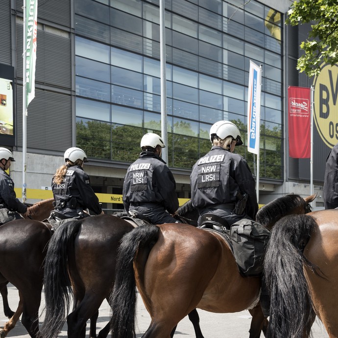 Sechs Menschen in Polizeiuniform mit weißen Helmen reiten nebeneinander von der Kamera fort. Im Hintergrund ist das Dortmunder Fußballstadion. (öffnet vergrößerte Bildansicht)