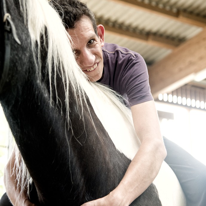 Ein Teilnehmer einer Reittherapiestunde sitzt auf einem schwarz-weißen Pferd und umarmt den Hals des Tieres. Er lächelt in die Kamera. (vergrößerte Bildansicht wird geöffnet)