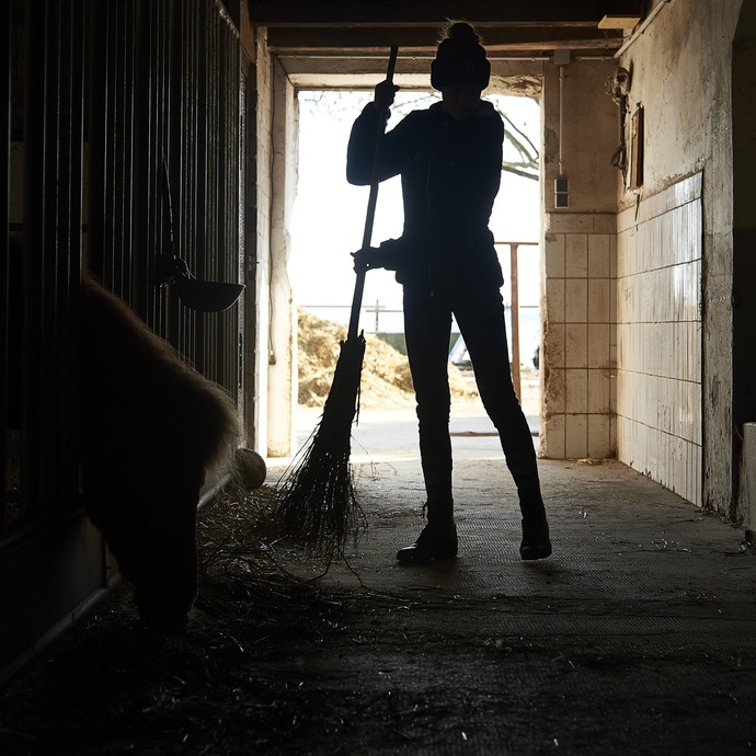 Ein Mädchen mit Mütze fegt im Gegenlicht eine Stallgasse. Man sieht nur ihre Silhouette. (vergrößerte Bildansicht wird geöffnet)