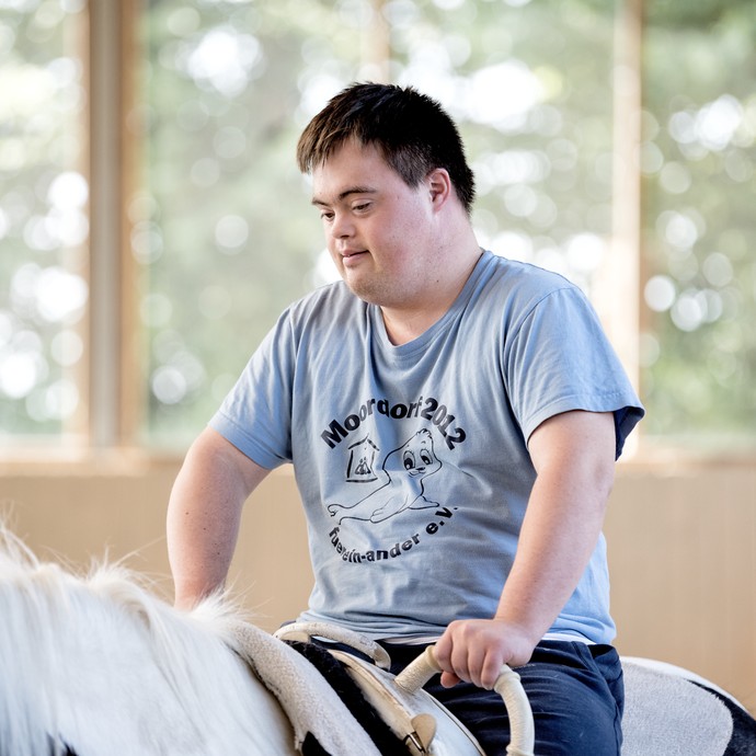 Ein Teilnehmer einer Reittherapiestunde sitzt auf einem schwarz-weißen Pferd. Er lächelt und hält sich an den Griffen des Sattels fest. (vergrößerte Bildansicht wird geöffnet)
