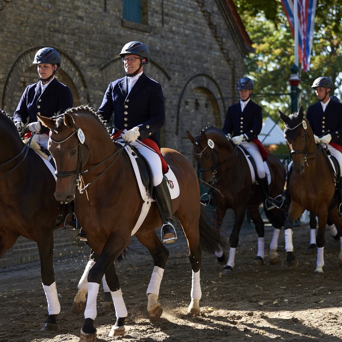 Fünf festlich gekleidete Menschen reiten auf braunen Pferde mit weißen Bandagen und verzierten Mähnen über einen Sandweg. (vergrößerte Bildansicht wird geöffnet)