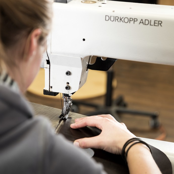 Blick über die Schulter einer Sattlerin, die an einer Nähmaschine arbeitet. Sie näht Lederstücke zusammen, auf der Maschine steht Dürkopp Adler. (vergrößerte Bildansicht wird geöffnet)