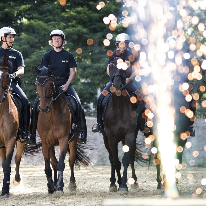 Mehrere Polizistinnen und Polizisten reiten nebeneinander auf braunen Pferden auf die Kamera zu. Im Vordergrund sprühen Funken. (vergrößerte Bildansicht wird geöffnet)