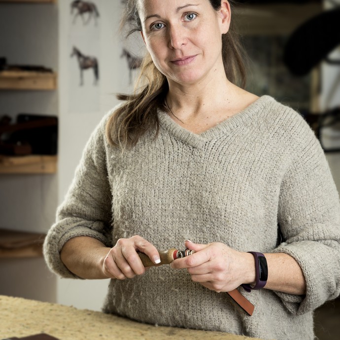 Eine Frau mit hellem Pullover blickt freundlich in die Kamera. Sie bearbeitet kleine Lederlaschen an einem Holztisch. (vergrößerte Bildansicht wird geöffnet)