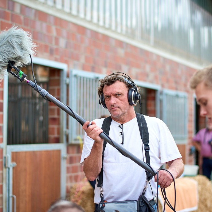 Ein Mann trägt ein Mikrofon an einer langen Stange, Kabel und eine Tasche. Er trägt Kopfhörer und guckt konzentriert. (öffnet vergrößerte Bildansicht)