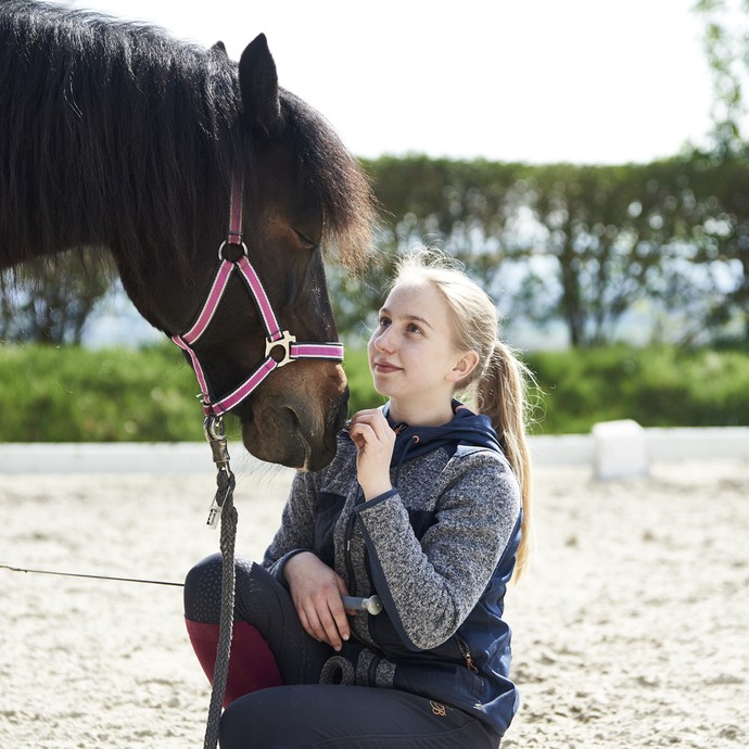 Ein blondes Mädchen kniet im Sand und schaut zu einem Pferd, welches den Kopf zu ihr hinab neigt. Sie berührt das Tier an der Nase und lächelt. (vergrößerte Bildansicht wird geöffnet)