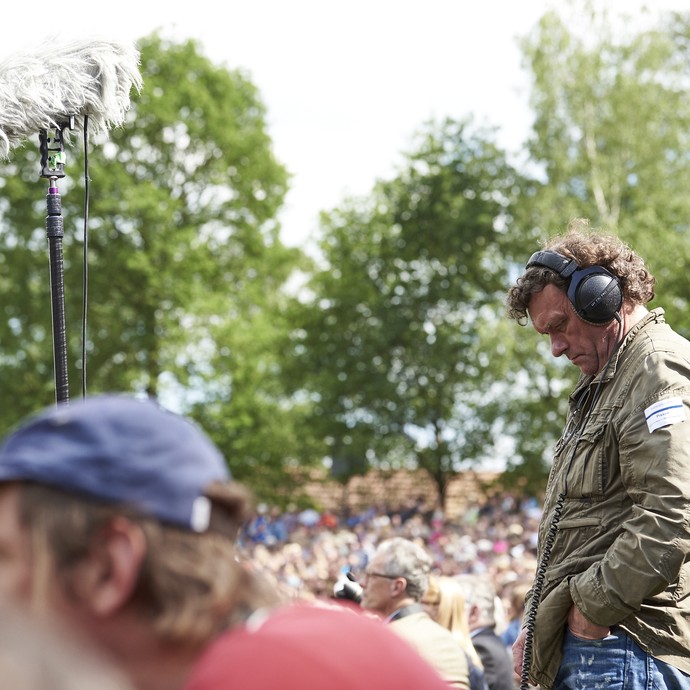 Ein Mann mit Kopfhörern steht in einer Menschenmasse. Vor ihm steht ein Mikrofon. Er scheint genau zuzuhören. (öffnet vergrößerte Bildansicht)
