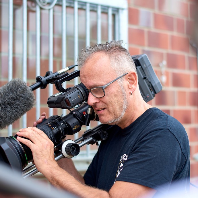 Ein Mann trägt eine große Filmkamera auf der Schulter. Er blickt durch den Sucher. Er macht einen Film. (vergrößerte Bildansicht wird geöffnet)
