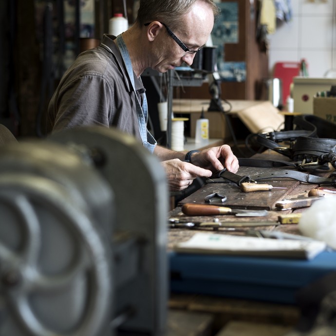 Ein Mitarbeiter einer Gestütswerkstatt sitzt an seinem Arbeitsplatz. Er bearbeitet eine Lederschnalle. Vor ihm liegen Werkzeuge auf dem Tisch. (öffnet vergrößerte Bildansicht)