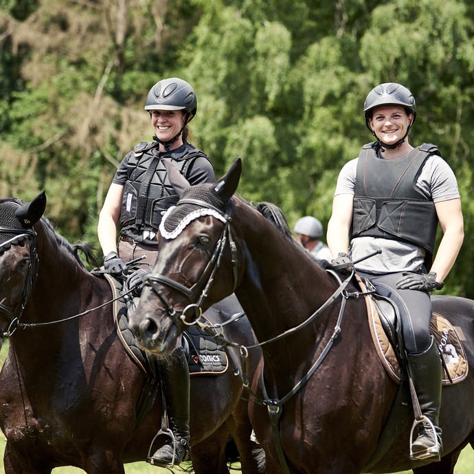Zwei Vielseitigkeitsreiterinnen auf dunklen Pferden sitzend lachen in die Kamera. Sie tragen Reithelme und Schutzwesten. (öffnet vergrößerte Bildansicht)