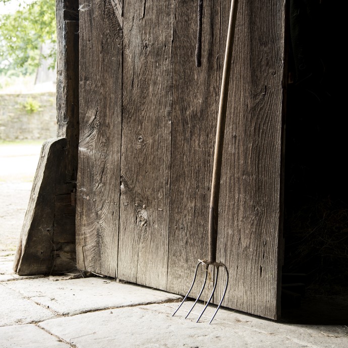 An einem Scheunentor aus Holz lehnt eine Mistgabel. Auf dem Boden liegen Natursteinplatten. (öffnet vergrößerte Bildansicht)