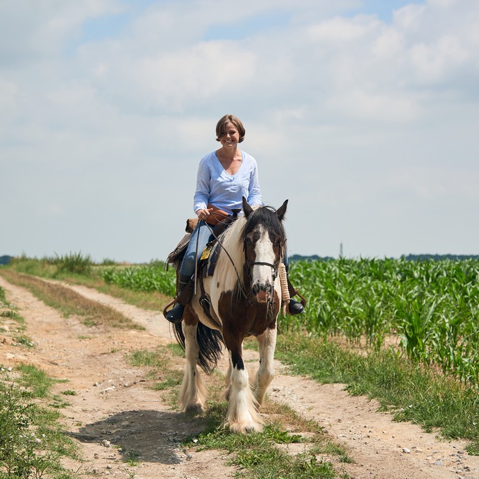 Eine Frau reitet einen Feldweg entlang. Das Pferd ist schwarz-weiß gefleckt. Die Frau lacht. (vergrößerte Bildansicht wird geöffnet)