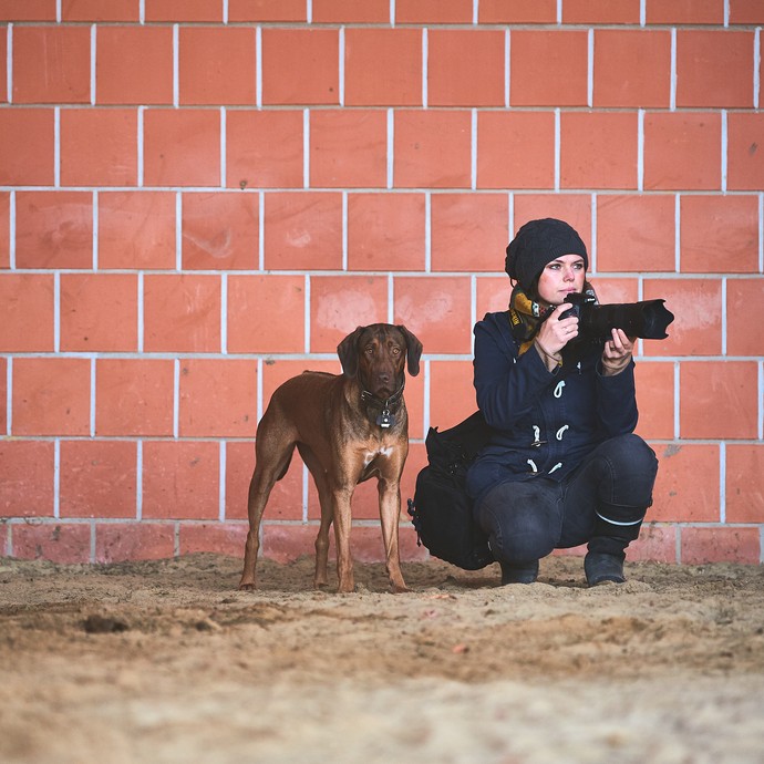 Eine Frau mit Kamera hockt auf dem Boden. Sie setzt an zum Fotografieren. Neben ihr steht ein Hund der in die gleiche Richtung blickt. (vergrößerte Bildansicht wird geöffnet)