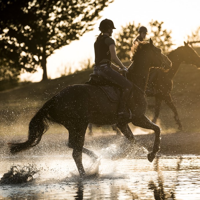 Zwei Reiterinnen galoppieren bei tief stehender Abendsonne mit ihren Pferden durch einen Wassergraben. (öffnet vergrößerte Bildansicht)